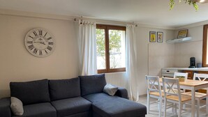 A living room with a dark grey sectional sofa, a small grey chest of drawers, a dining table with white chairs, a window with white curtains, and a wall clock. - Le Cottage du Brask (La Motte-du-Caire)