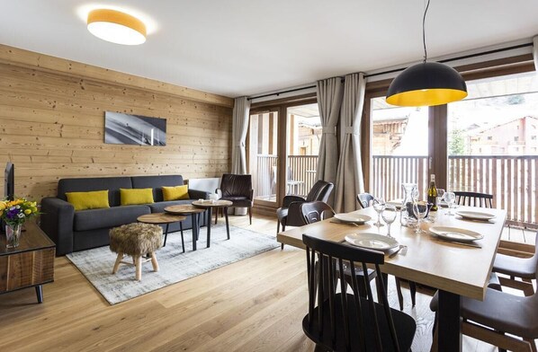 Een moderne woonkamer met een houten vloer, een bank met gele kussens, een eettafel gedekt voor vier personen, en een groot raam met gordijnen.