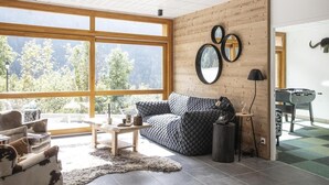 A modern living room with a large window, wooden walls, a sofa, a coffee table, and a foosball table. - Wifi, view mountain, sauna, hammam, terrace, balcony, parking, ski locker, 98-106m², Vaujany (Vaujany)