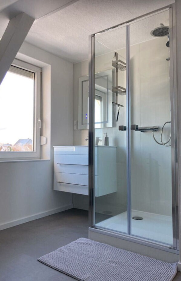 Une salle de bain moderne dotée d’une douche à parois de verre, d’un meuble-lavabo blanc et d’une grande fenêtre qui laisse entrer la lumière naturelle.