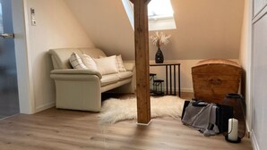 Un dormitorio tipo loft acogedor, con techo inclinado, un sofá color beige, un cofre de madera y una mesita con un florero.