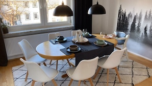 Un comedor con una mesa redonda, sillas blancas, un mantel oscuro y un mural de un bosque en la pared.