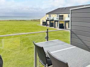 A balcony with a view of the sea, a grassy lawn, and a building with multiple doors and windows. - 5 Person Holiday Home in Lokken (Lokken)