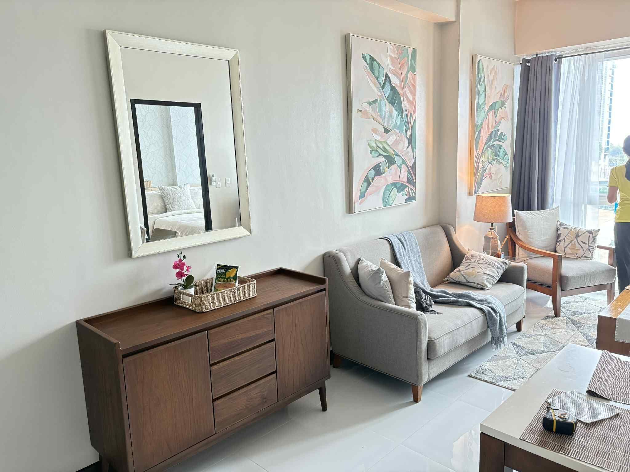 Una sala de estar moderna con un sofá gris, un mueble de madera y un espejo grande. Hay cuadros abstractos en la pared y una ventana con cortinas.