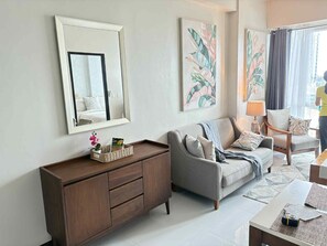 A modern living room with a grey sofa, a wooden cabinet, and a large mirror. There are abstract paintings on the wall and a window with curtains. - Mactan Newton Condo with Pool Access (Lapu-Lapu)