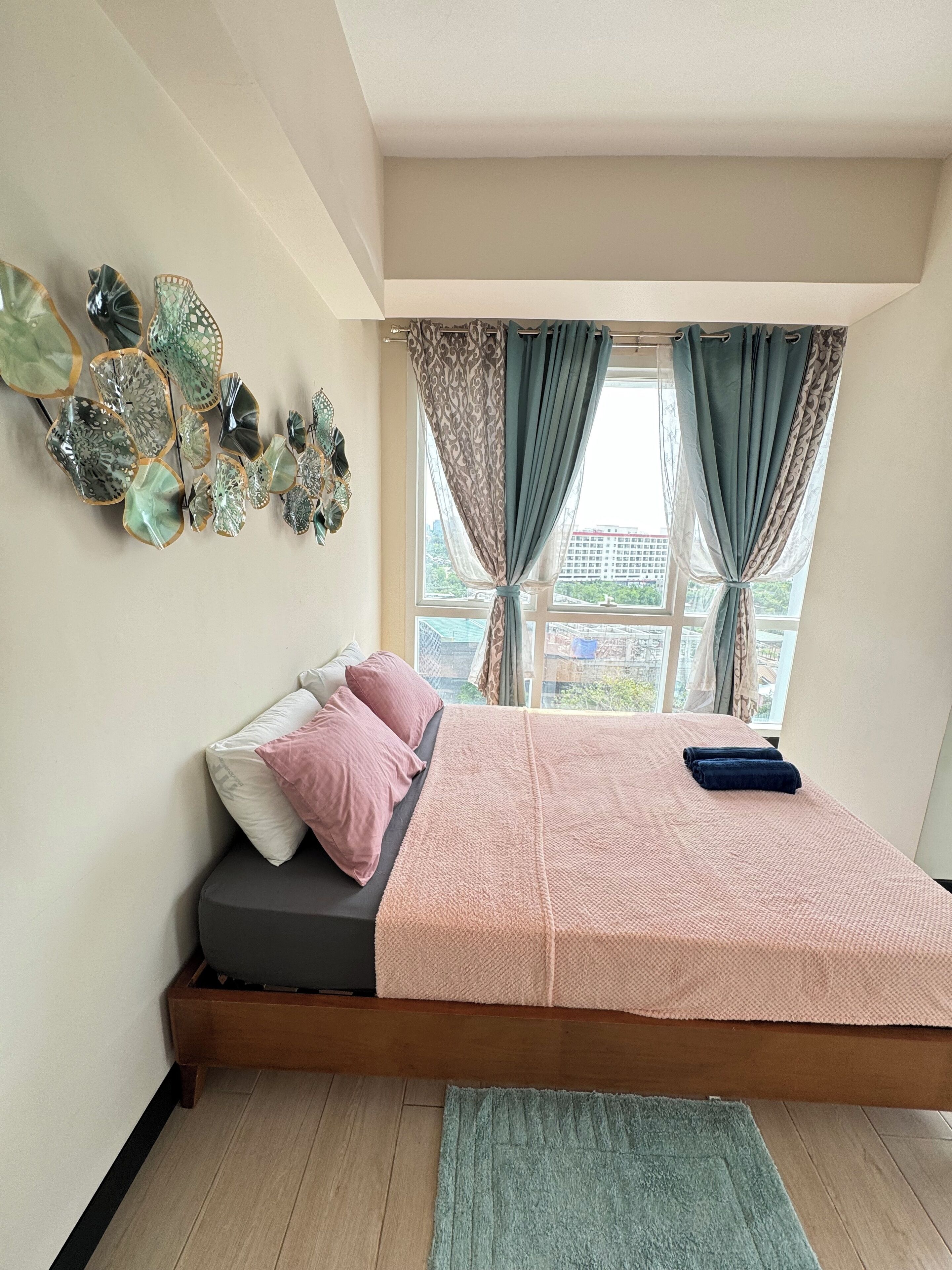 Un dormitorio con cama, almohadas, mesita de noche, ventana con cortinas y un adorno en la pared.
