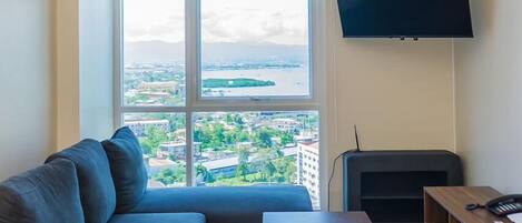 Una sala de estar con un sofá, una mesa de centro, un televisor de pantalla plana y vistas a un paisaje urbano a través de una ventana con persianas.