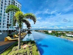 A rooftop infinity pool with yellow loungers, surrounded by palm trees and a view of a cityscape. - Mactan Newton Condo with Pool Access (Lapu-Lapu)