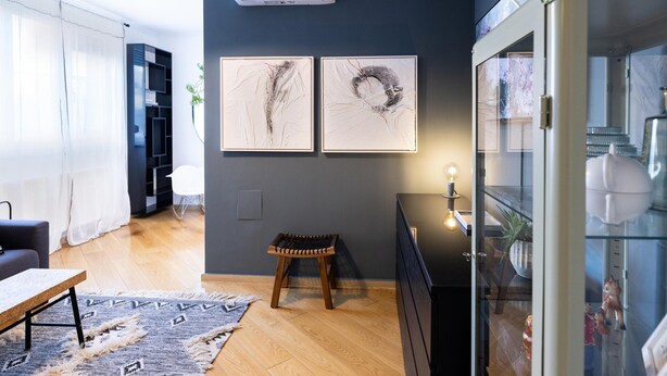 A modern living room with a sofa, a coffee table, a rug, and a small stool. There are two framed artworks on the wall and a glass cabinet with decorative items.