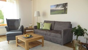 A living room with a grey sofa, a wooden coffee table with fruit, a grey armchair, a lamp, and a painting of a tree. - Holiday Apartment Borkowski (Welzheim)