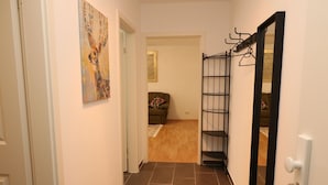 Un couloir avec un sol carrelé, une porte ornée d’un porte-clés, une étagère en métal noir et un tableau accroché au mur.
