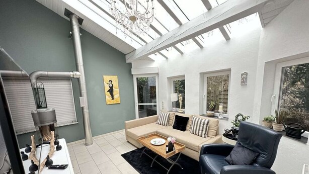 Una sala de estar moderna con un sofá, un sillón y una mesa de comedor. Se ve un ventanal con vistas a árboles y una araña.