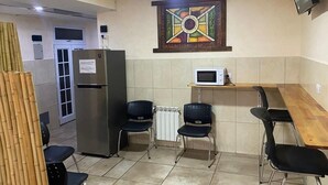 Une petite cuisine équipée d’un réfrigérateur, d’un four à micro-ondes et de deux chaises. Il y a une fenêtre avec des stores et un vitrail décoratif au-dessus du four à micro-ondes.