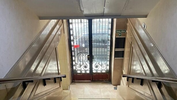 Un escalier menant à une porte surmontée d’une grille métallique, une pancarte indiquant « SORTIE » et un luminaire au plafond.