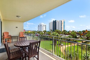 A balcony with a wooden table and chairs, a view of a cityscape with tall buildings, and a well-maintained lawn with palm trees. - Sterling Shores 307 (Destin)