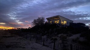 A beach house with a unique roof design, situated on a sandy beach with a wooden fence in the foreground. - Atypical, set on the beach with direct access, swimming pool, barbecue, pets allowed (Marseillan)