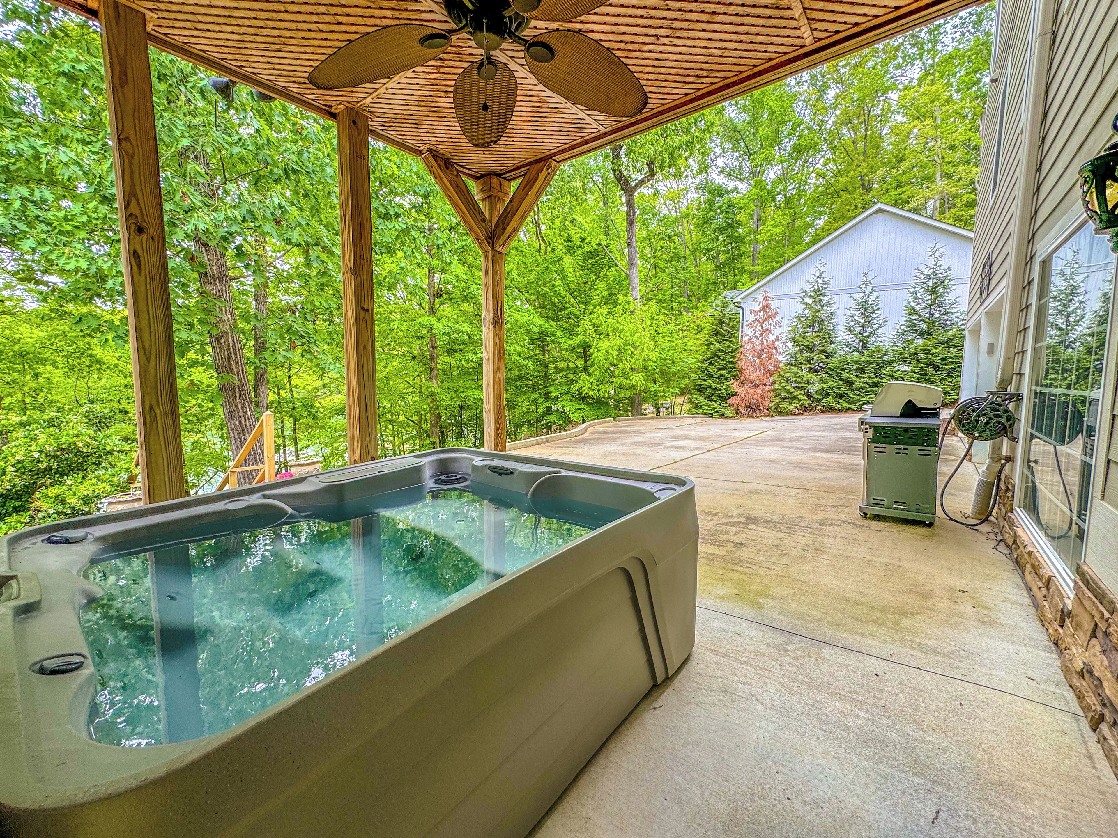 A hot tub under a wooden pergola with a ceiling fan, surrounded by lush greenery.