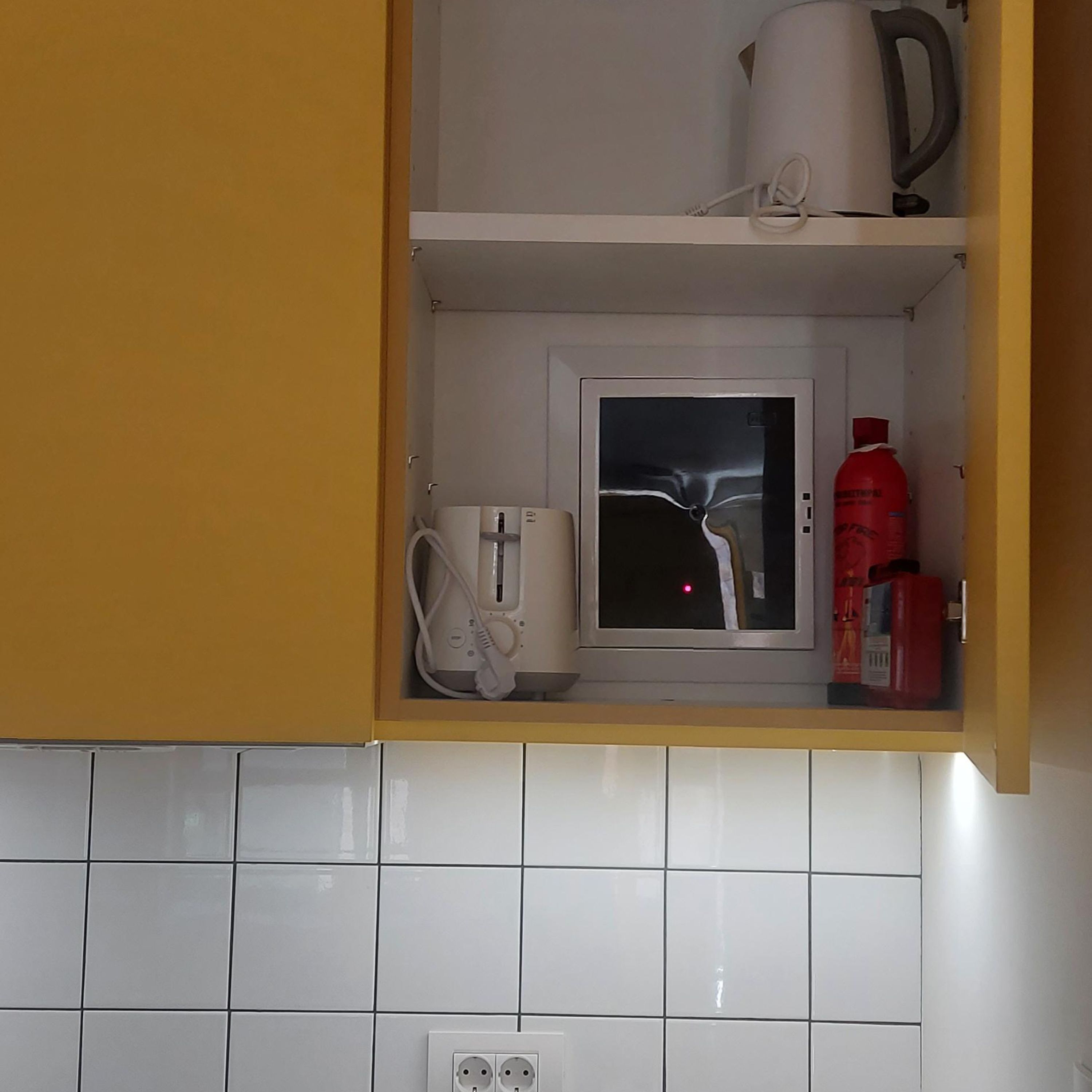 A kitchen with a microwave, kettle, and red fire extinguisher on a shelf.