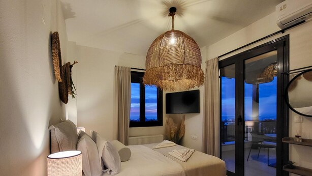Quarto moderno com cama, luminária, televisão e vista da cidade ao entardecer.