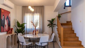 Uma área de refeições com uma mesa redonda, cadeiras e plantas. Há uma escada com degraus de madeira e corrimão.