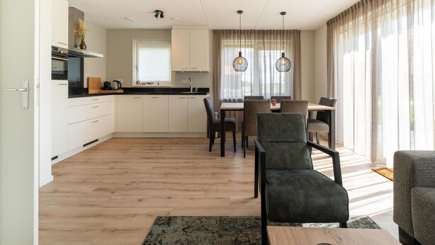 Een moderne keuken met witte kasten, een zwart aanrecht en een raam boven de gootsteen. Er is een zithoek met een tafel en stoelen, en een bankje op de voorgrond.