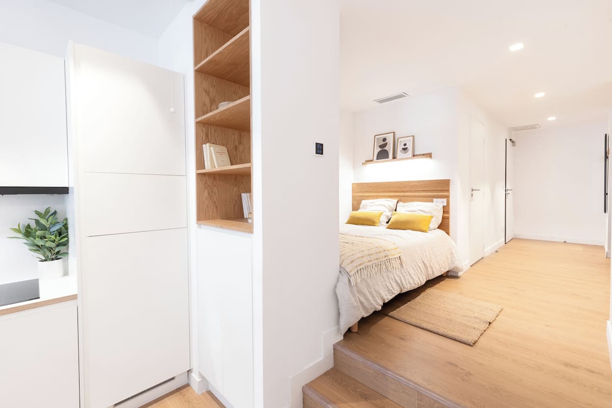 Une chambre à coucher moderne avec une tête de lit en bois, un lit avec une literie blanche et jaune, une petite table de chevet et une étagère intégrée remplie de livres.