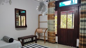 Une chambre d’hôtel comprenant un lit, une table de chevet, une chaise, une étagère en bois, une fenêtre avec des rideaux et une porte.