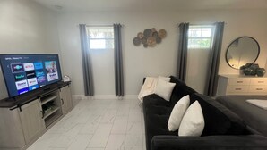 Un salón con televisor de pantalla plana mostrando diferentes servicios de streaming, un sofá de color oscuro con almohadas, un mueble blanco y un espejo grande en la pared.