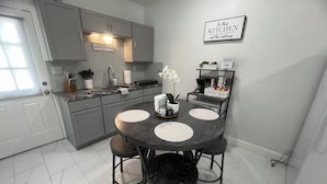 Una cocina moderna con armarios grises, una encimera de mármol y una mesa de comedor con cuatro sillas. Hay una barra de café con un letrero, una planta en maceta y un cartel que dice 'En esta cocina contamos los recuerdos, no las calorías'.