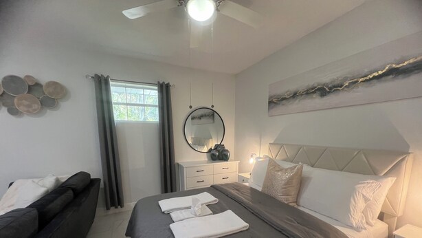 Habitación de hotel con cama, sofá, mesita de noche, espejo y ventana con cortinas.