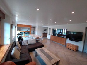 A modern living room with a sofa, coffee table, and a dining area with a table and chairs. A kitchen with a TV and cabinets is visible in the background.