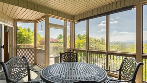 Une terrasse couverte avec une table ronde et des chaises, de grandes fenêtres offrant une vue sur les arbres et les montagnes.