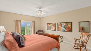 Une chambre avec un grand lit, un ventilateur de plafond, une fenêtre avec des rideaux et deux tableaux accrochés au mur.