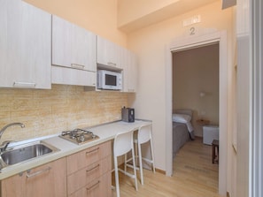 A compact kitchen with a microwave, a gas stove, and a sink. There is a small dining area with a white chair and a bed in the adjacent room.