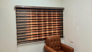 Una habitación con un ventanal grande cubierto por persianas escocesas, un sillón de cuero marrón y una pequeña mesa de madera.