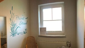 A room with a wicker chair, a small table with a cup, a window with a sign, and a wall with a decorative plant and anchor design. - Dat Onnen's Hus - Burhave 3 (Butjadingen)