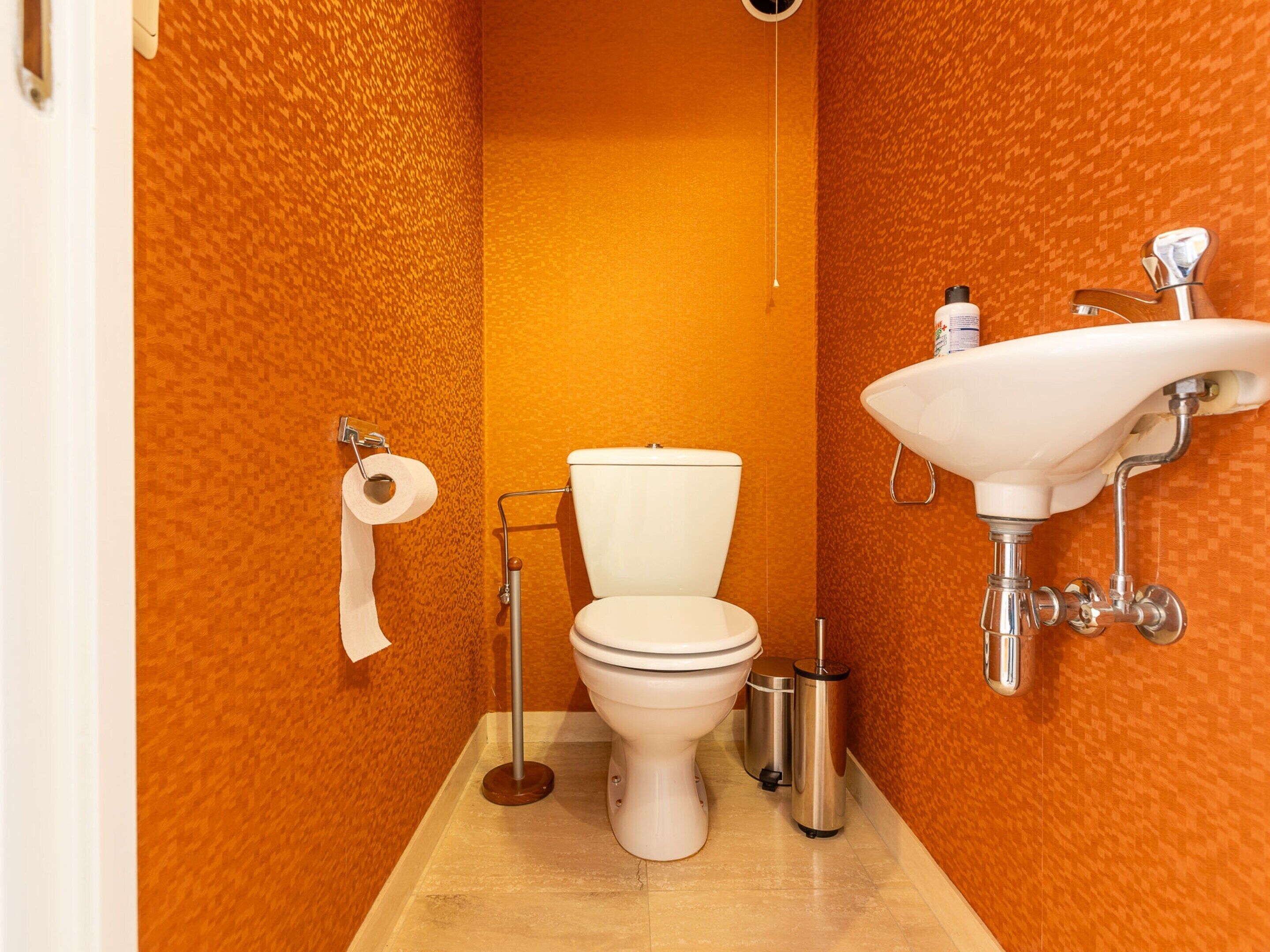 Ein Badezimmer mit orangefarbener Wandstruktur, einer weißen Toilette, einem wandmontierten Waschbecken und einem Mülleimer.
