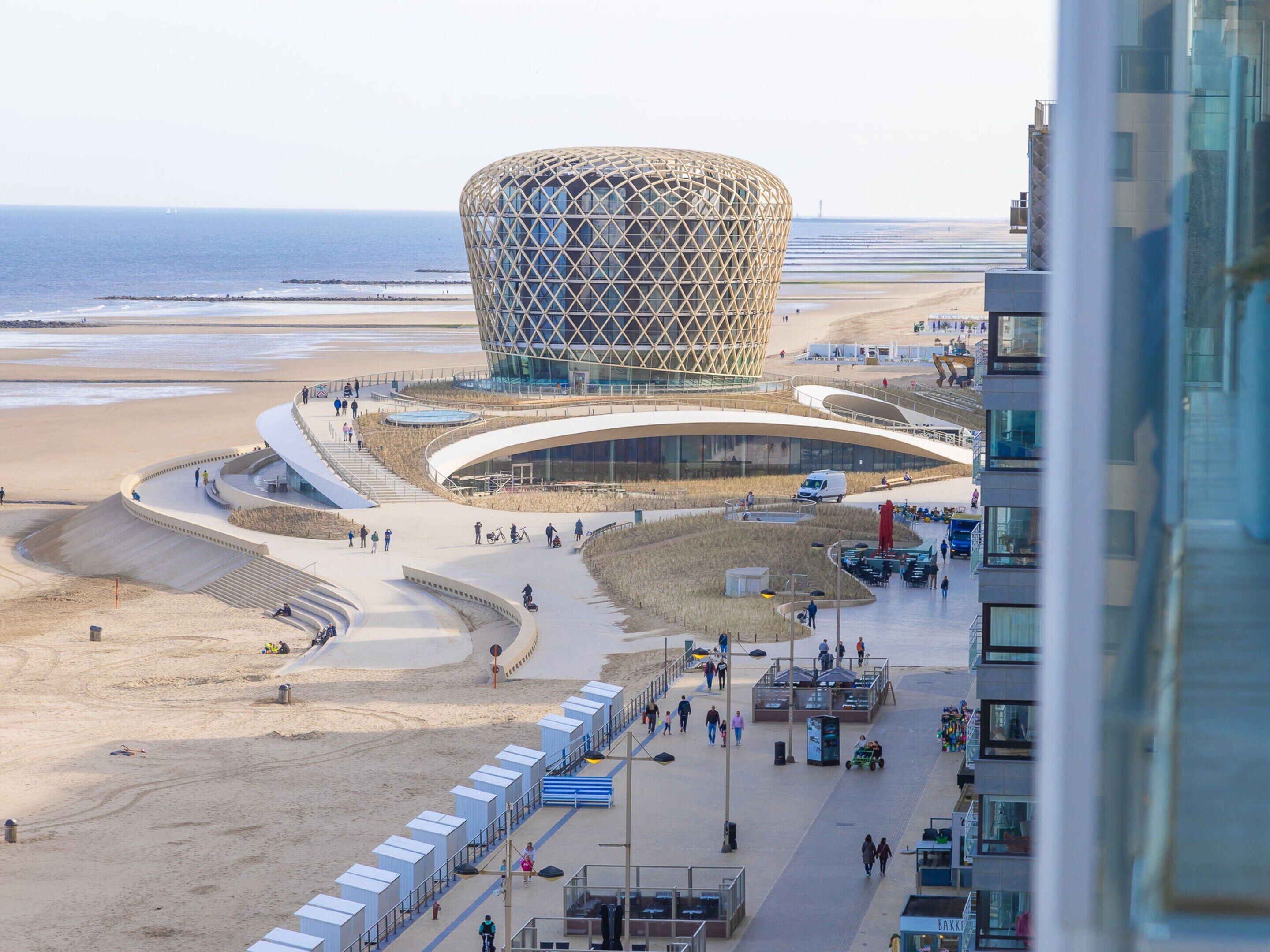 A modern building with a distinctive geometric design, situated on a beachfront promenade with a sandy beach and the ocean in the background.