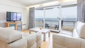 Un salon moderne doté d’un téléviseur à écran plat, de canapés beige, d’une table basse et d’un balcon offrant une vue sur la mer.