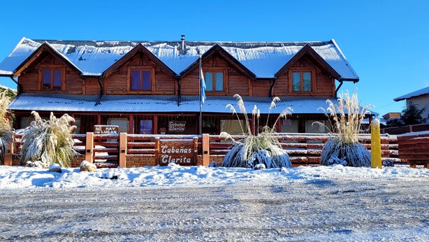 A wooden building with a corrugated metal roof, a sign reading "Cabanas Clarita," and a wooden fence in front. - Cabañas Clarita (Dina Huapi)