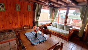A wooden dining area with a table, chairs, and a bench. There are curtains on the windows, and a small table with a vase of flowers. - Cabañas Clarita (Dina Huapi)