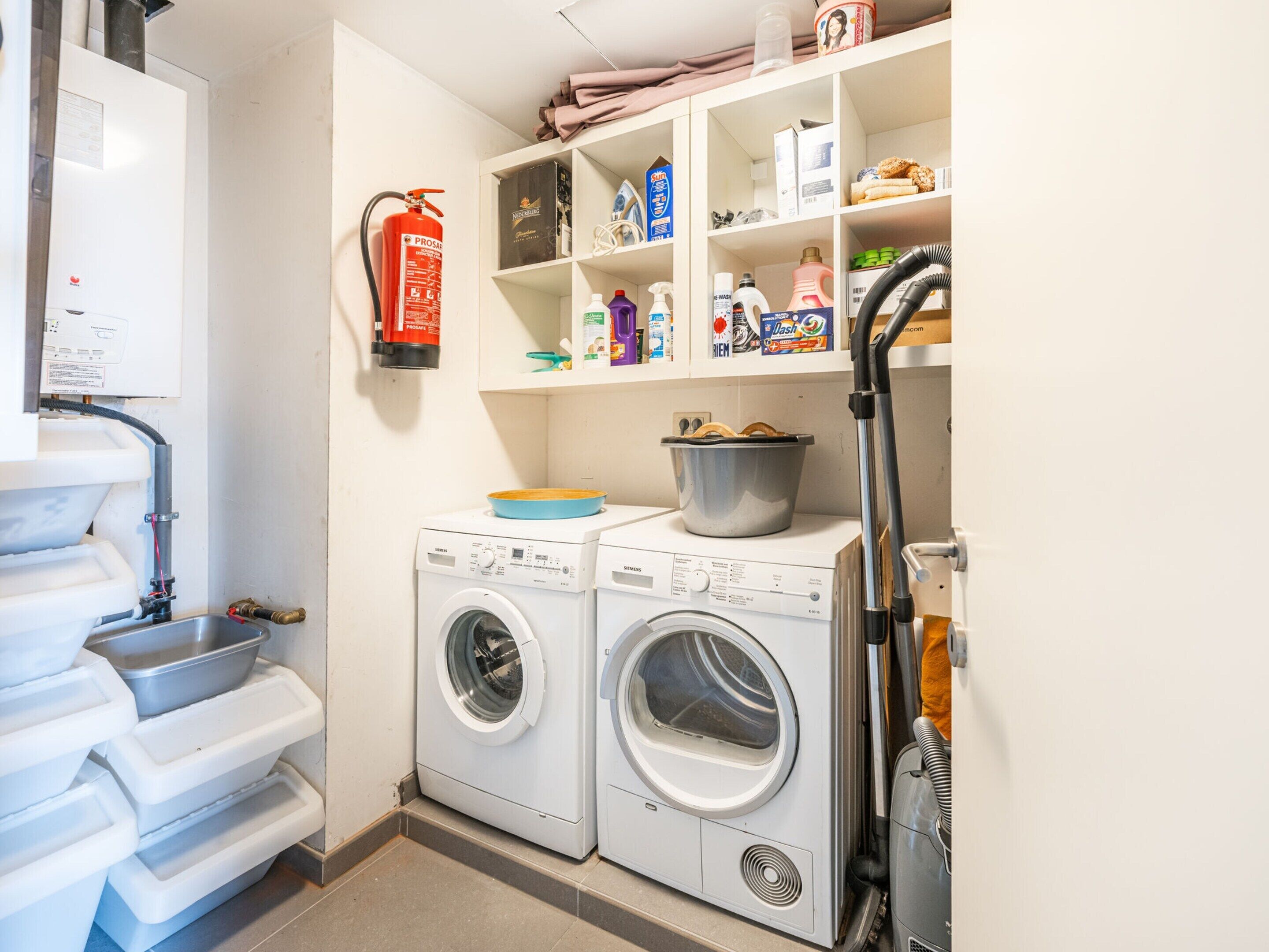 A compact laundry room with two front-loading washing machines, a fire extinguisher, and a shelf with various cleaning supplies.