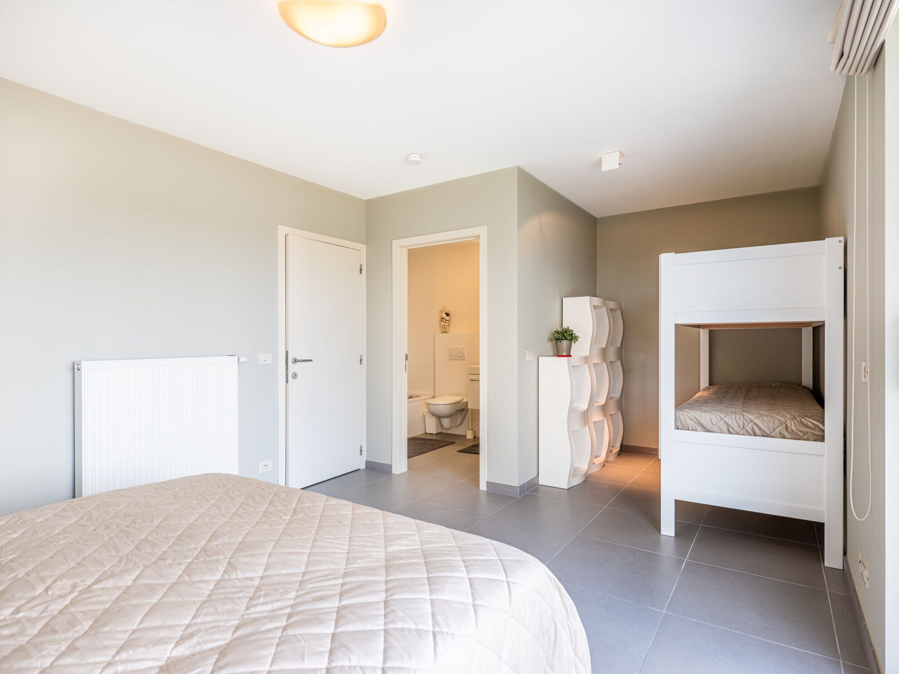 A bedroom with a large bed, a bunk bed, a radiator, and a bathroom visible through an open door.