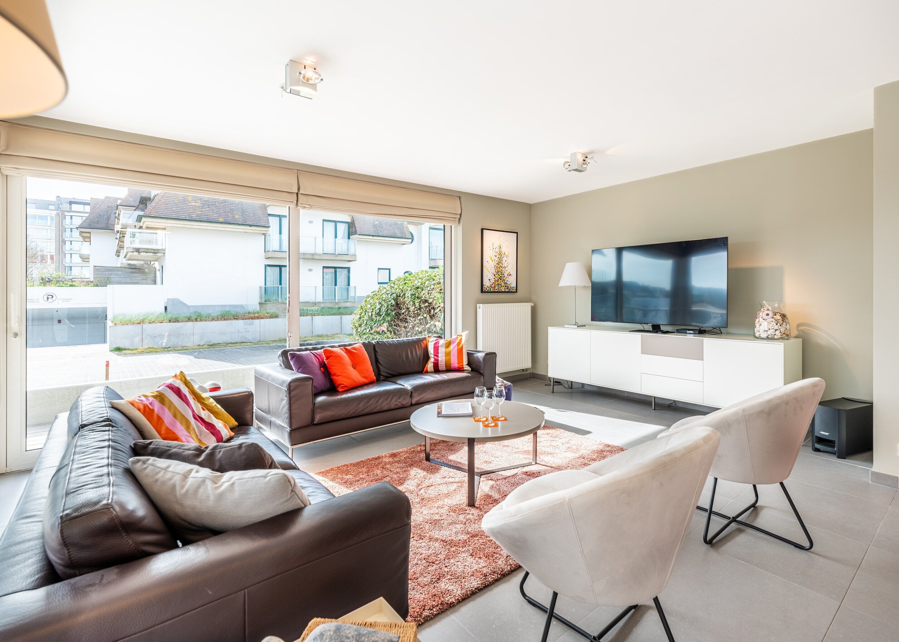 A modern living room with a sofa, coffee table, and TV, large windows, and a view of buildings outside.