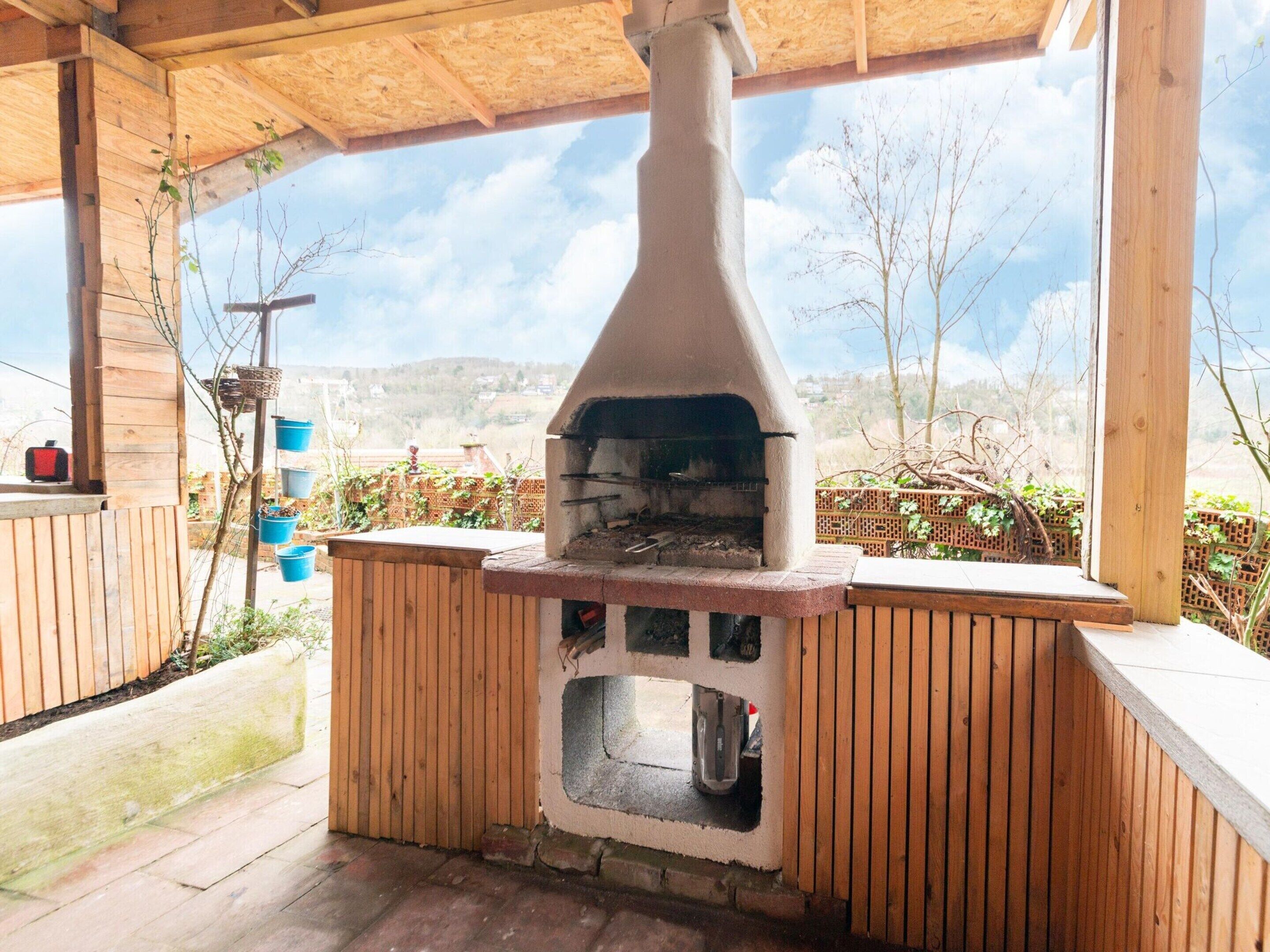 Estructura de madera al aire libre con un horno de leña integrado, rodeada de muros de madera y una vista despejada del paisaje.
