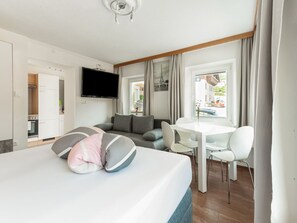 A modern hotel room with a bed, a sofa, a dining table with chairs, a TV, and a view of the street.