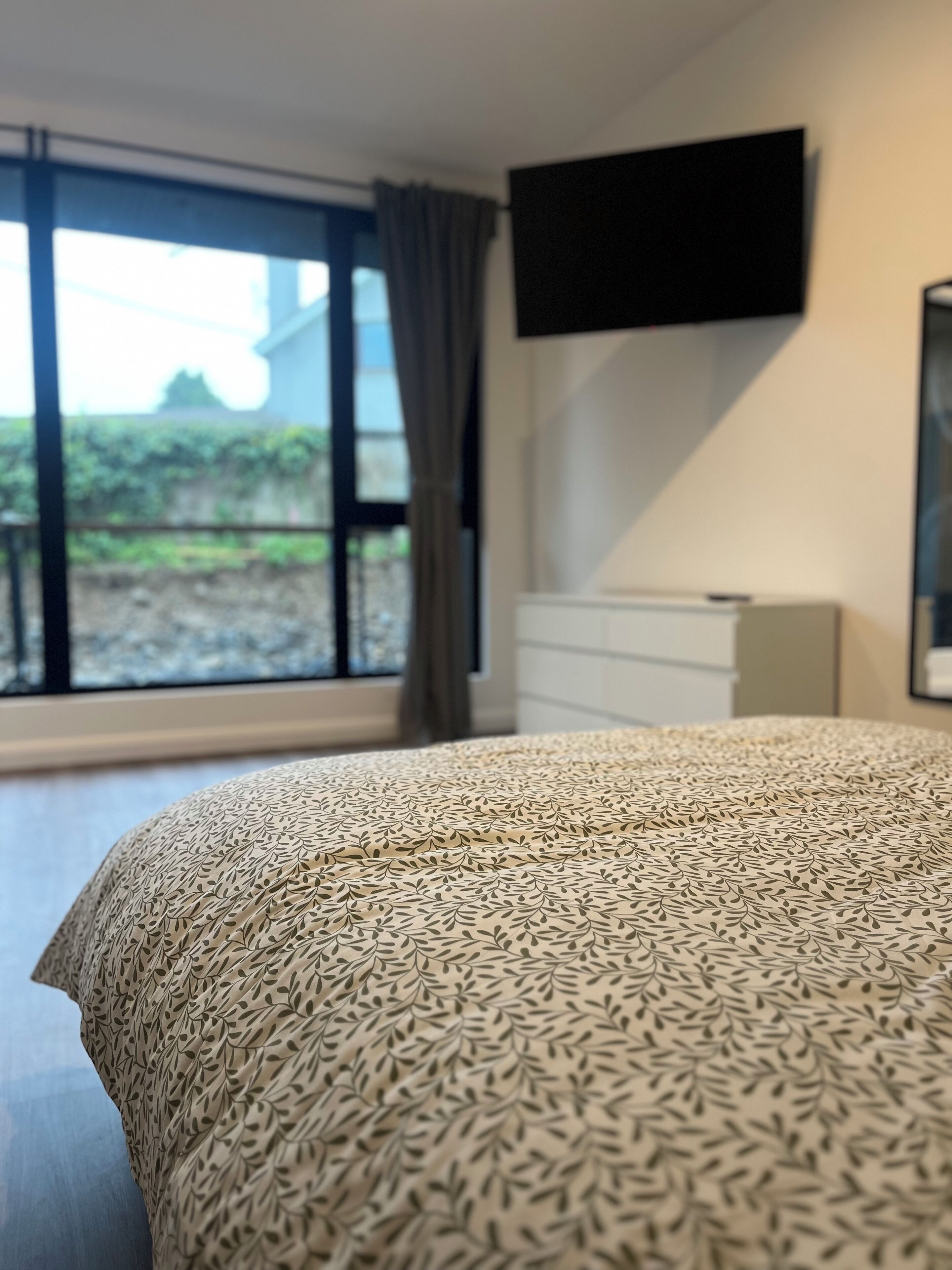 A bedroom with a large window, a bed with a patterned bedspread, a flat-screen TV mounted on the wall, and a dresser.