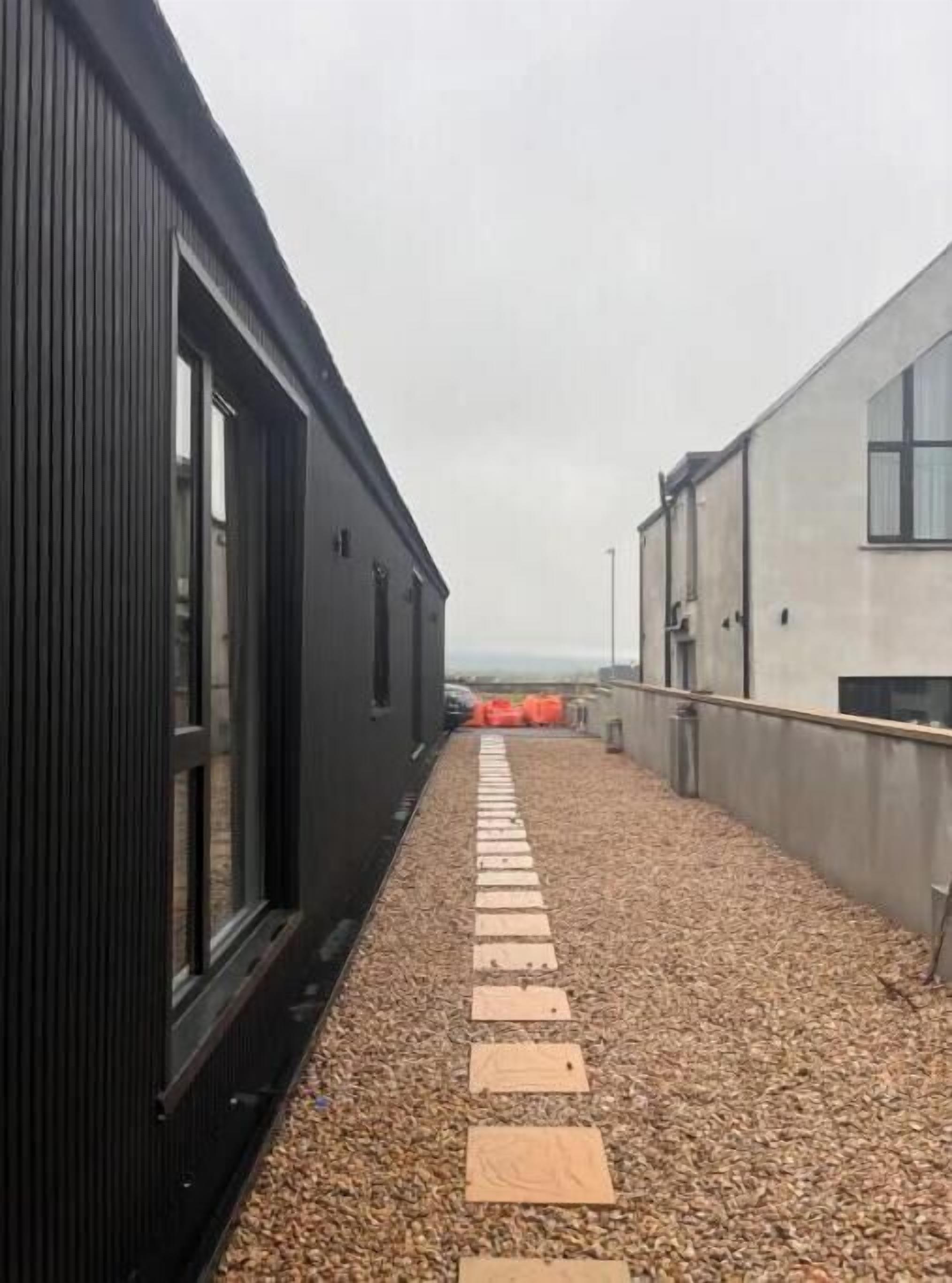 Un sentier étroit, agrémenté de marches en bois, mène à un bâtiment doté de grandes fenêtres.