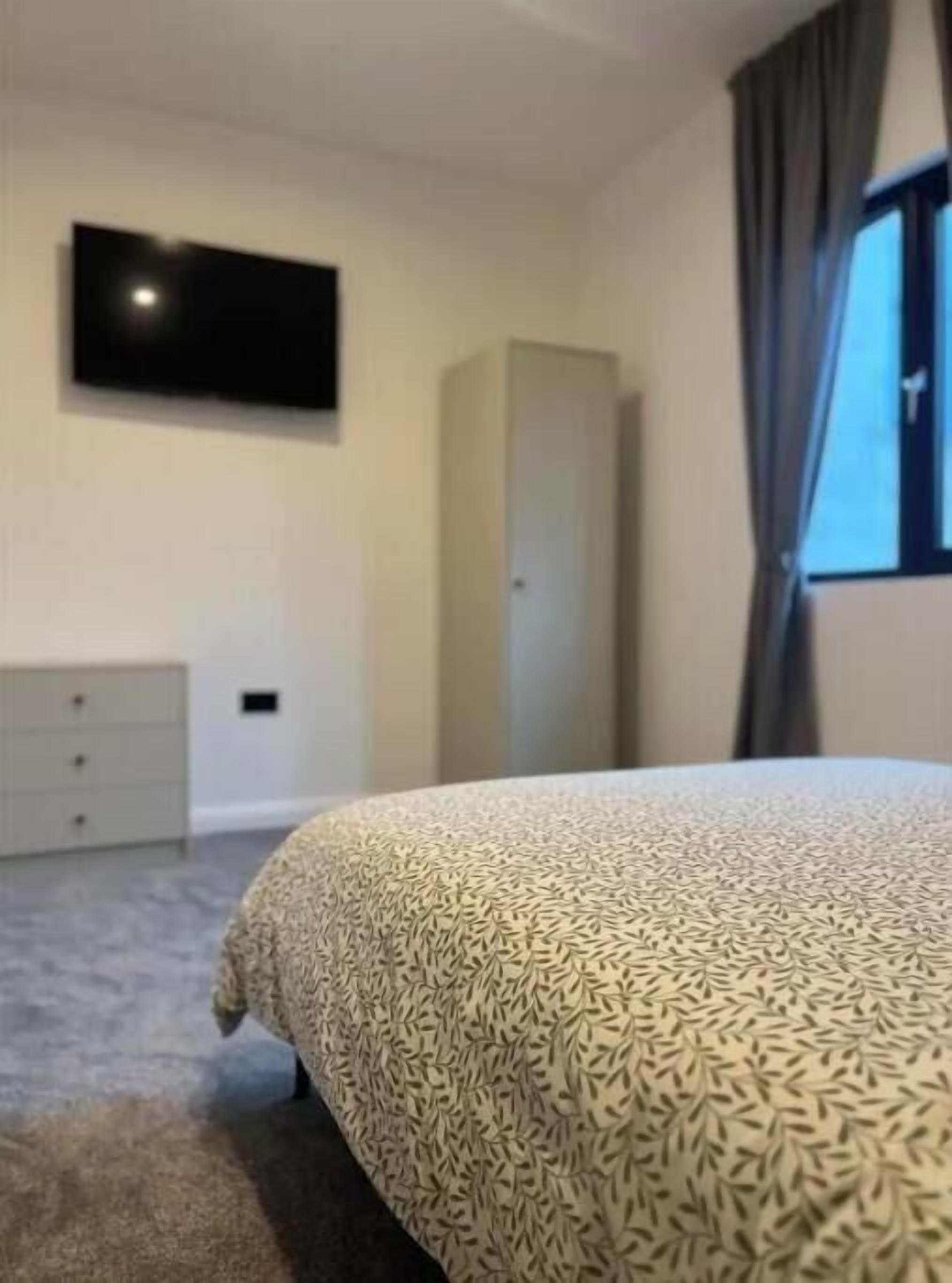 A bedroom with a bed, a television mounted on the wall, a wardrobe, and a window with curtains.
