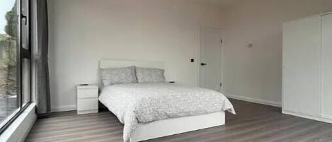 Ein modernes Schlafzimmer mit großem Fenster, einem Bett mit weißem Kopfteil, einem Nachttisch und einem Kleiderschrank.